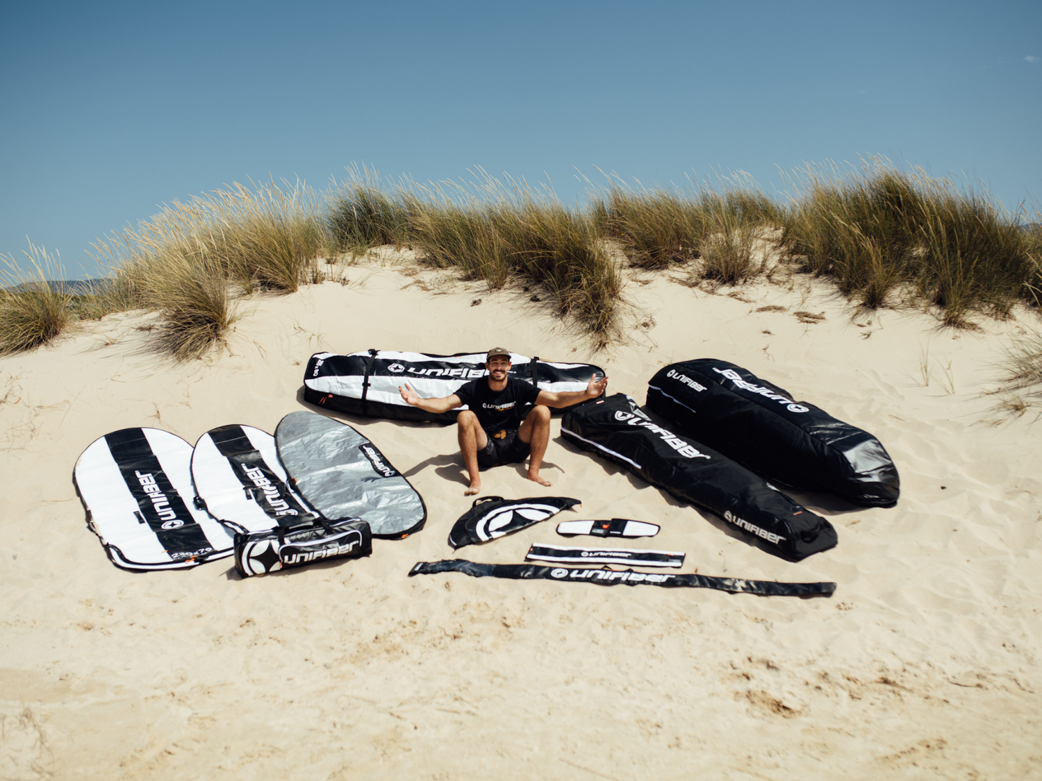 Unifiber windsurf bag solutions: Comprehensive display of protective bags for boards, masts and sails laid out on sandy dunes.