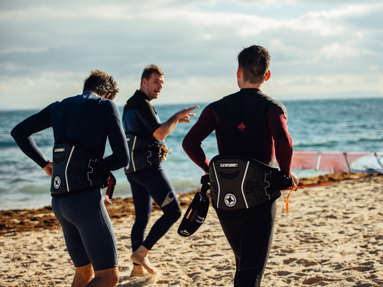 Three windsurfers wearing Unifiber harnesses walking along beach shoreline before entering water.