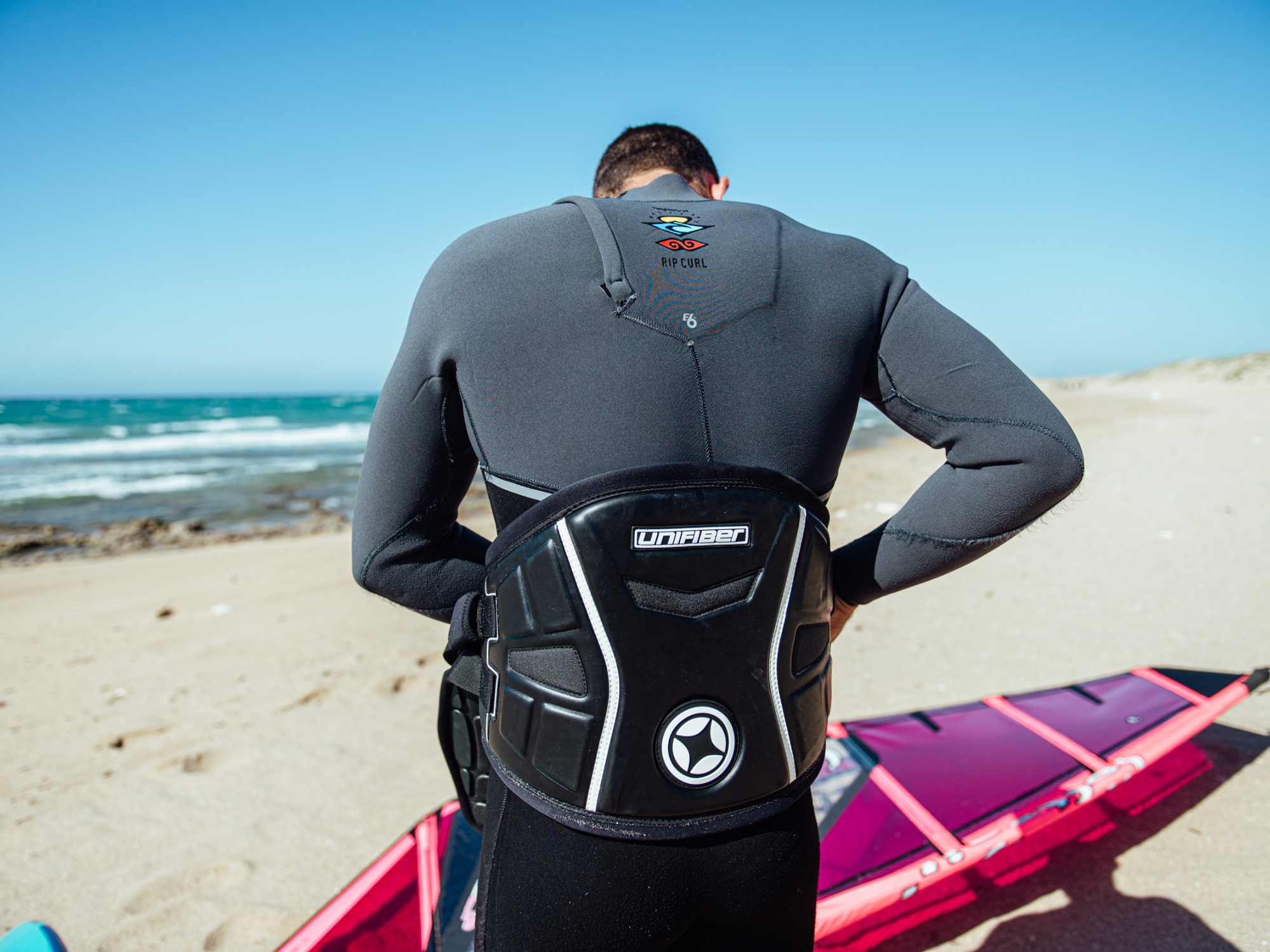 Unifiber windsurf harness design: Person wearing grey wetsuit with black harness on beach next to pink windsurf equipment