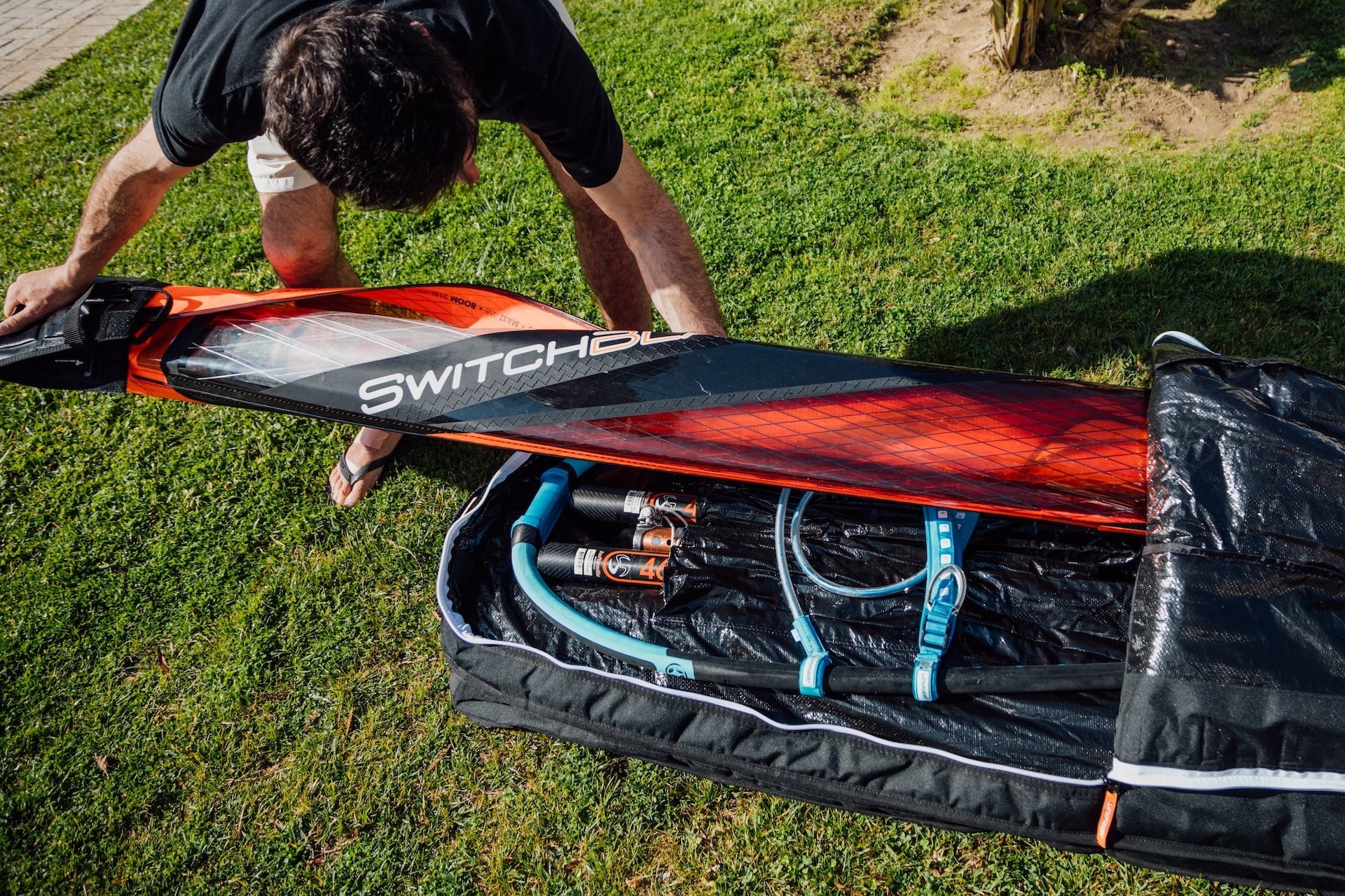 a windsurfer putting his sail into a windsurf quiver bag where we can a windsurf boom and a windsurf mast