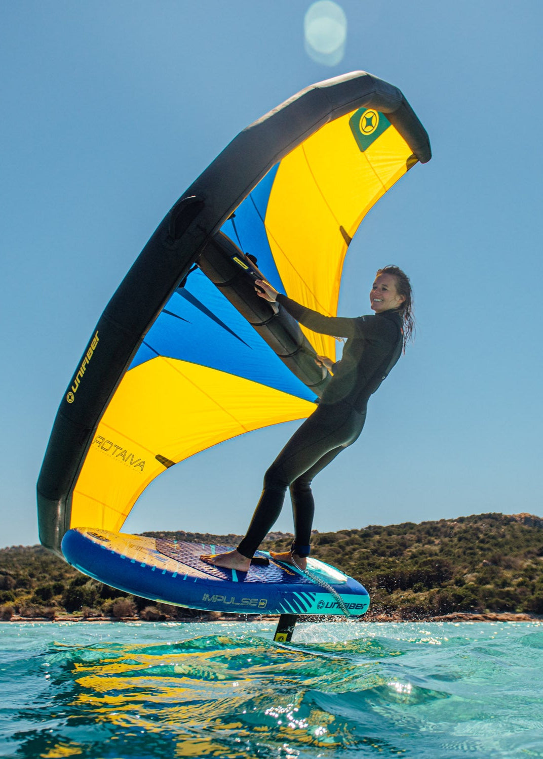 A rider riding a blue hydrofoil board, controlling a yellow and blue wing sail against a coastal backdrop.