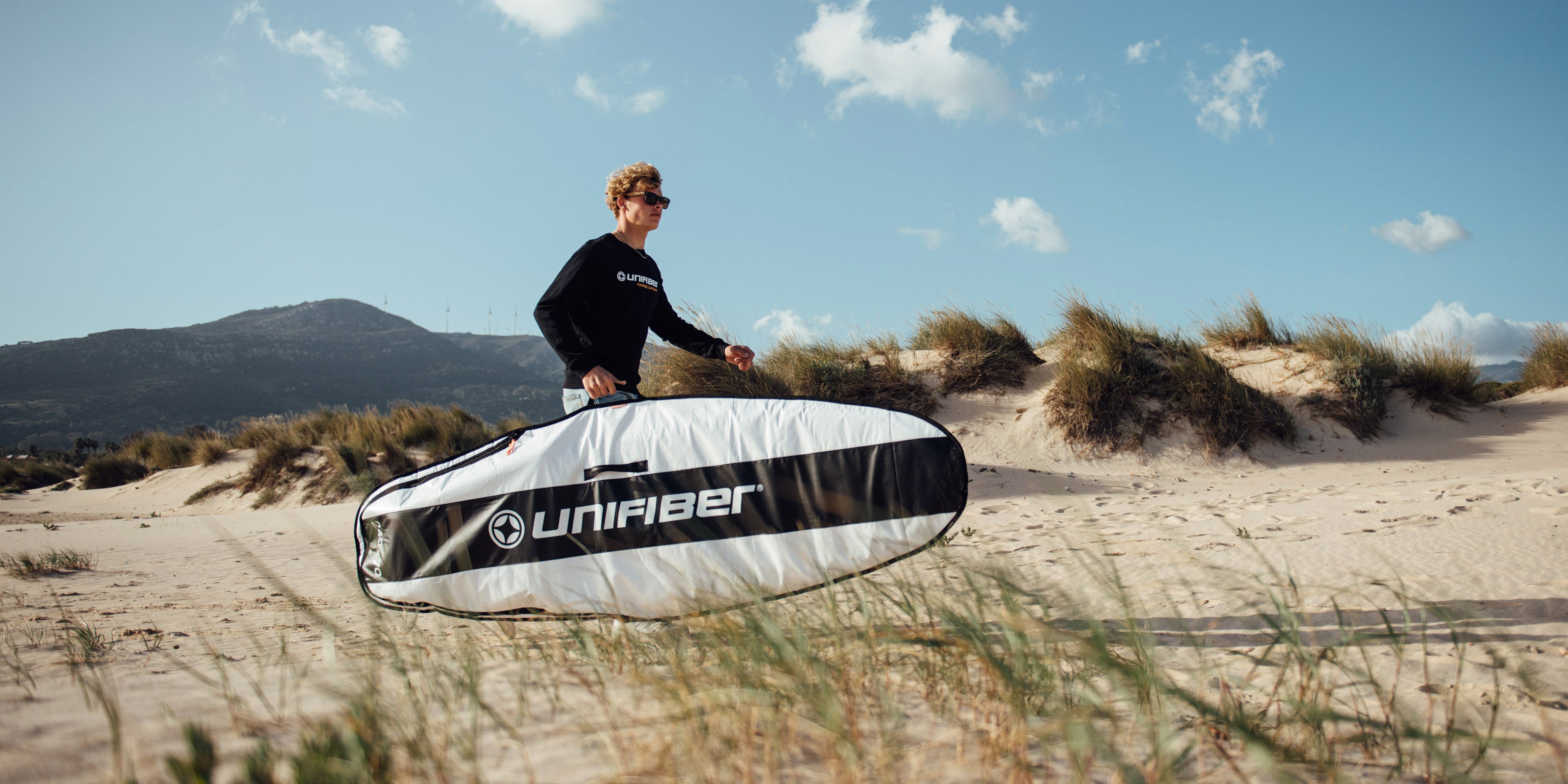 A person wearing sunglasses and a black Unifiber sweatshirt stands on a sand dune holding a white and black Unifiber board bag. The scenic background features beach grass, sand dunes, and mountains under a blue sky with scattered clouds