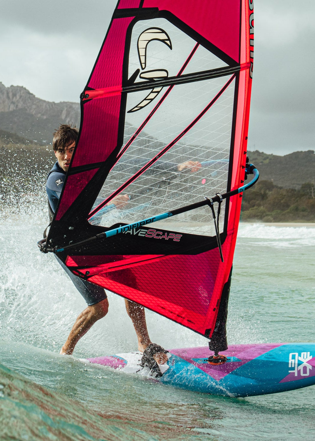 someone windsurfing using a Wave Scape sail in red and white colors. They're riding on a colorful board with blue and pink accents while navigating choppy waters. Mountains are visible in the background, and water is spraying as they maneuver across the surface. They're wearing what appears to be a dark wetsuit while handling the windsurfing equipment.