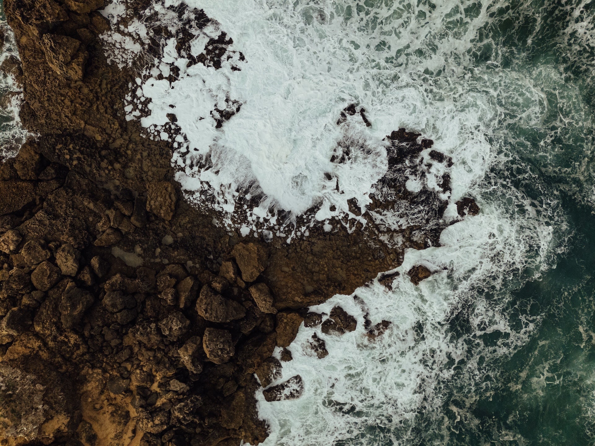 Aerial view of ocean waves creating white foam patterns as they crash against dark rocky coastline