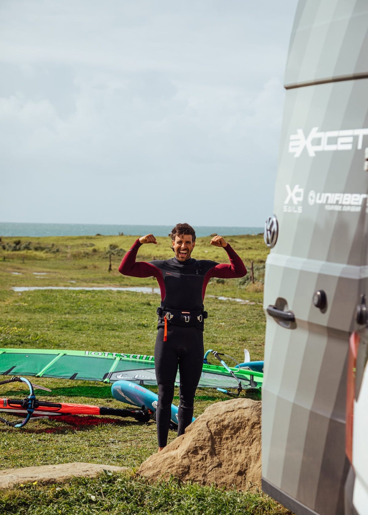Windsurfer in black wetsuit flexing his arms next to white windsurfing equipment on grassy terrain