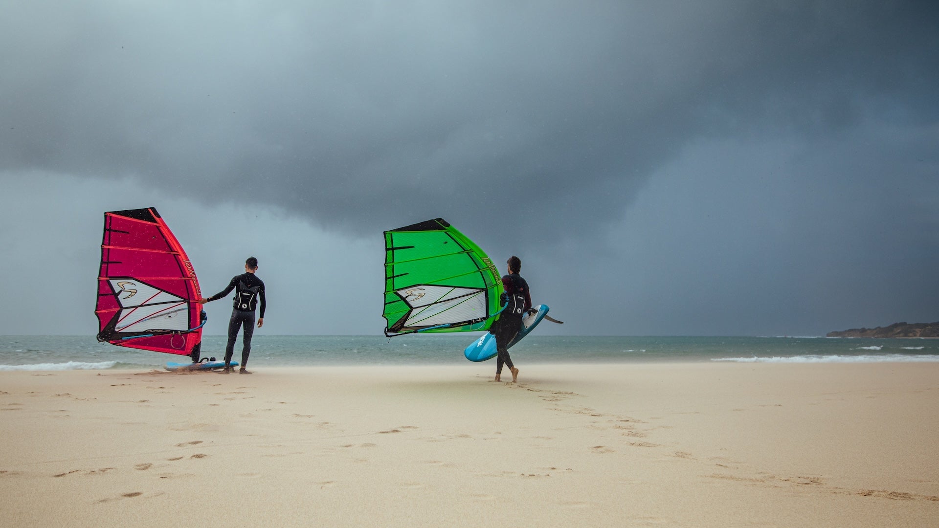 Two windsurfers in black wetsuits stand on a beach during overcast weather. One holds a red sail while the other carries a bright green sail. The ocean appears choppy under stormy gray skies, creating a moody atmosphere. Both riders are preparing their equipment on the sandy shore