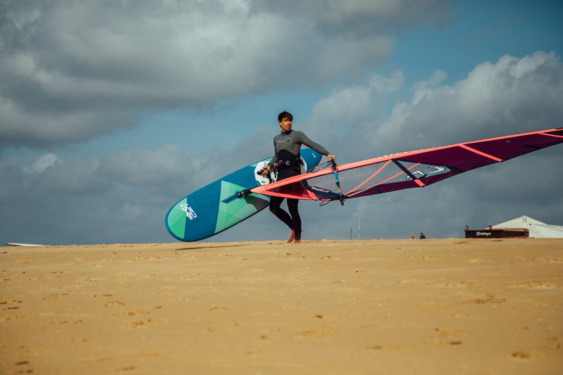 Windsurfer balancing windsurfing equipment on a sandy beach against dramatic cloudy skies