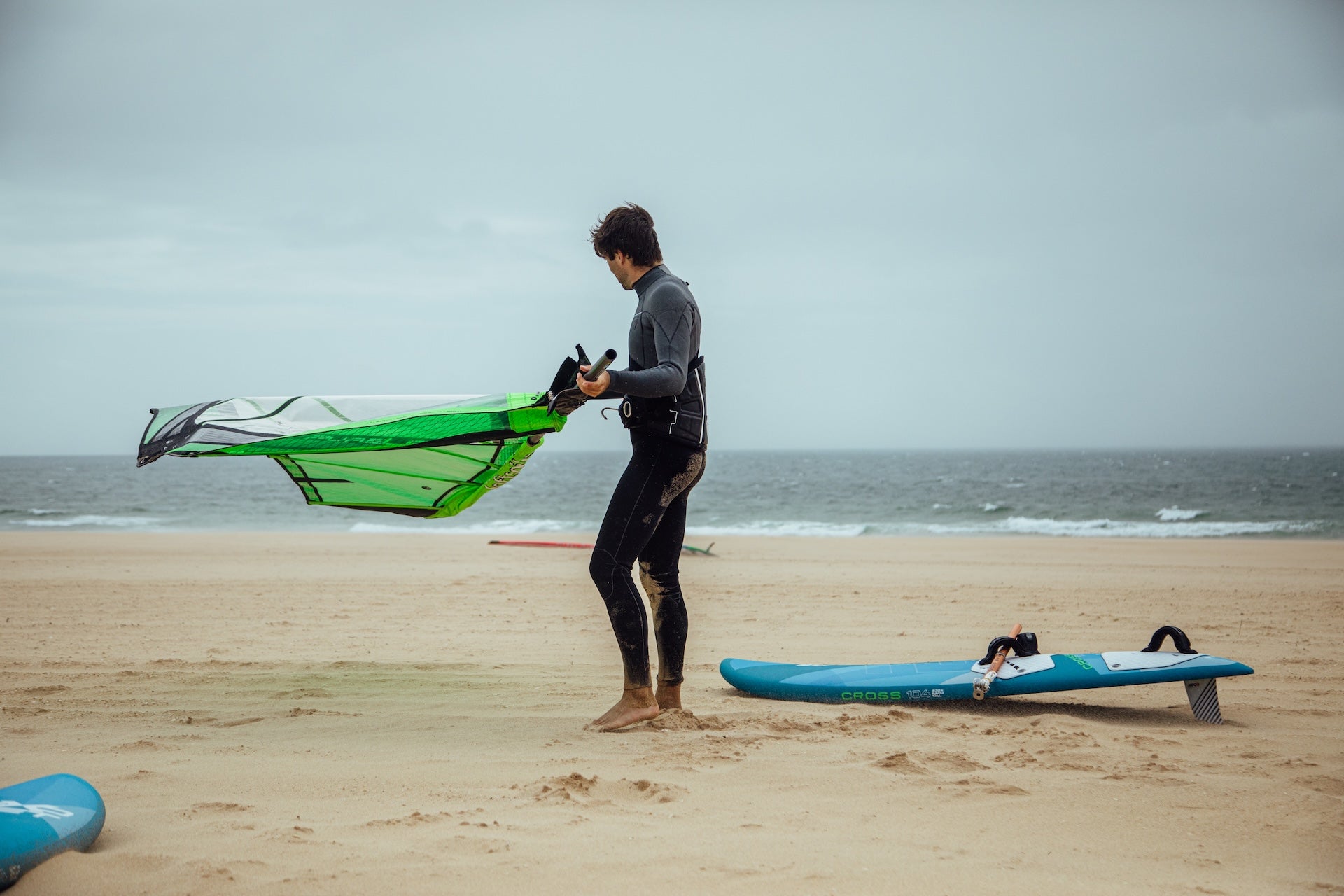 Windsurfer in grey wetsuit rigging a bright green sail on a sandy beach, with a blue exocet windsurf board lying nearby.