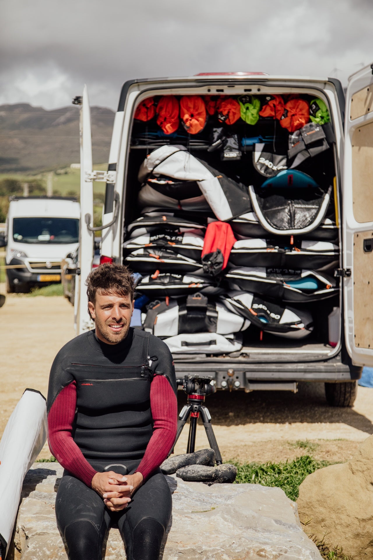 Smiling windsurfer in black and red wetsuit sitting beside an open van packed with multiple windsurf boards and equipment