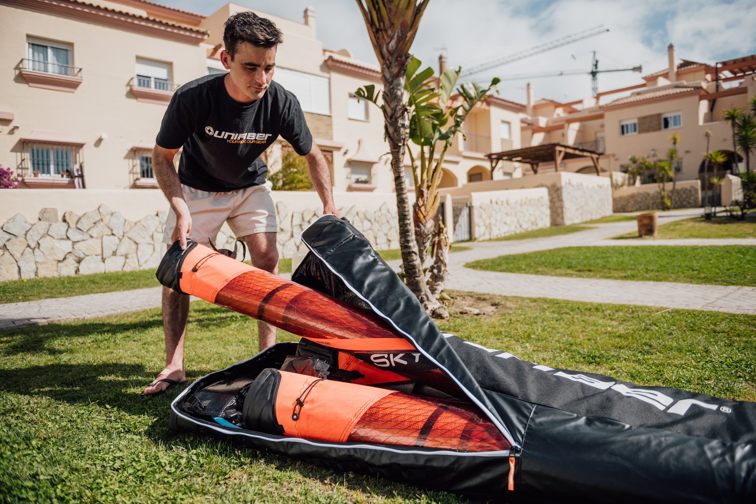 Person wearing a black Unifiber t-shirt and beige shorts organizing windsurf equipment into a black gear bag with red-orange sails, on a lawn in front of Mediterranean-style vacation homes.
