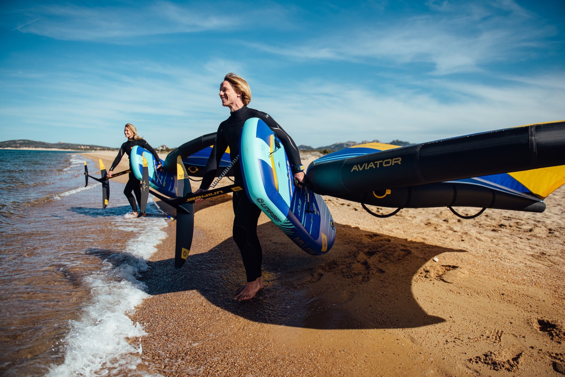 two riders on the beach with watersport equipment it a wingfoil set with a hydrofoil and inflatable board