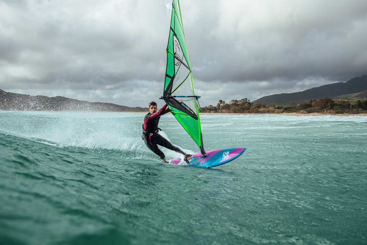 A windsurfer glides across choppy turquoise waters on a cloudy day, riding a pink and blue board with a green and black sail against a backdrop of misty mountains and gray skies
