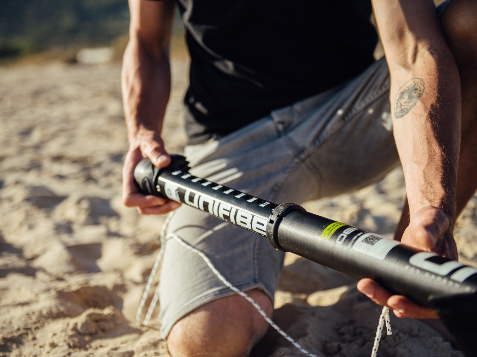 1. Close-up photo of hands adjusting rope on Unifiber windsurf mast extension on sandy beach.