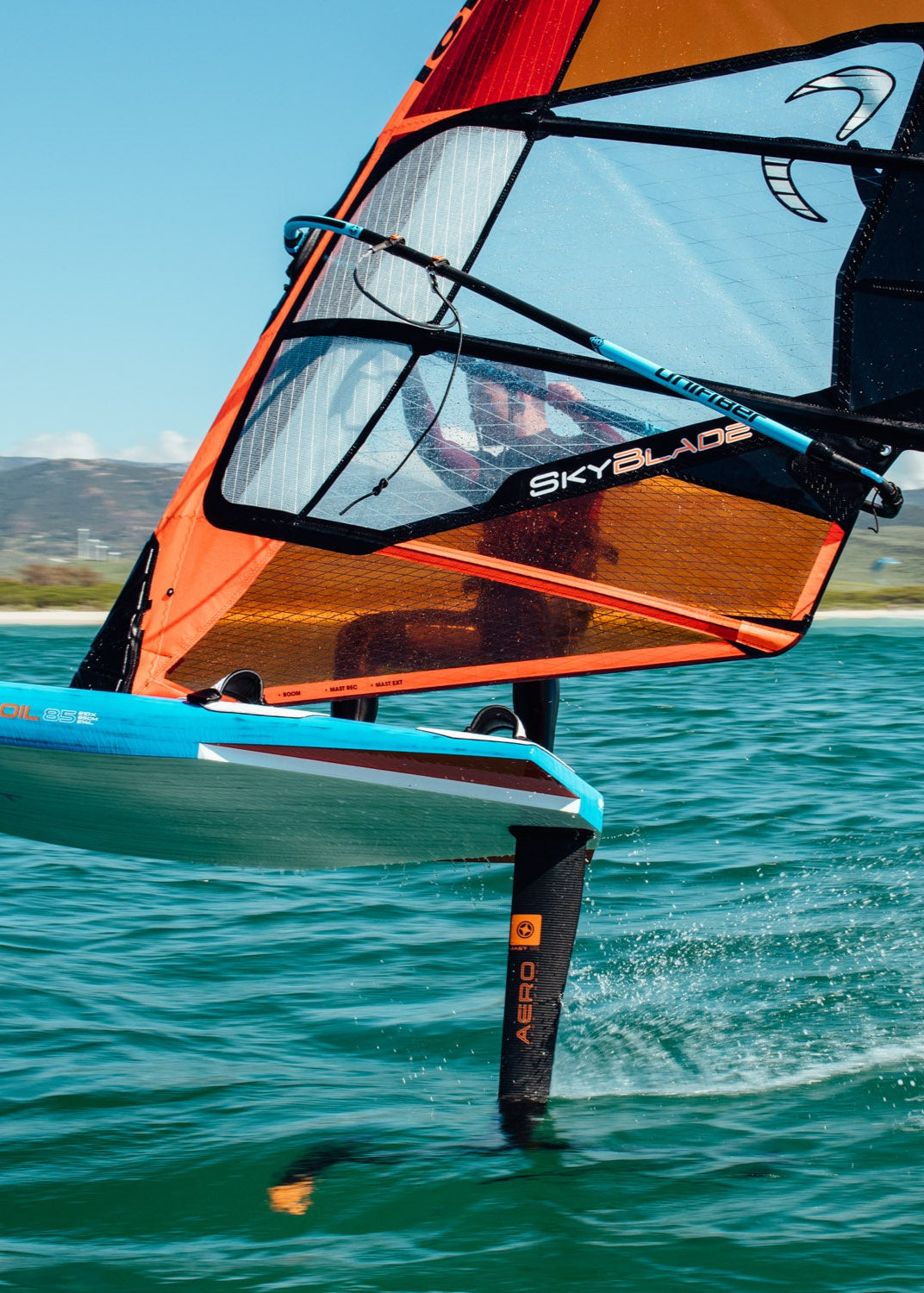 s a close-up of windsurfing equipment in action. The orange and black SkyBlade sail is visible along with a hydrofoil lifting the blue board above the water's surface. Mountains and a beach are visible in the background under clear blue skies.