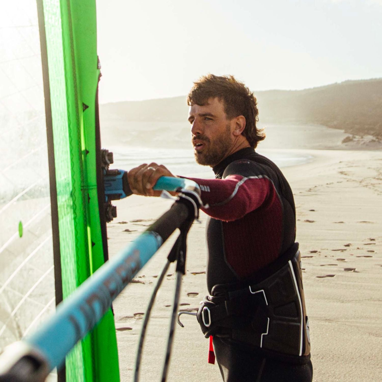 A person adjusting windsurfing equipment on a beach at sunset, wearing a black and burgundy wetsuit.
