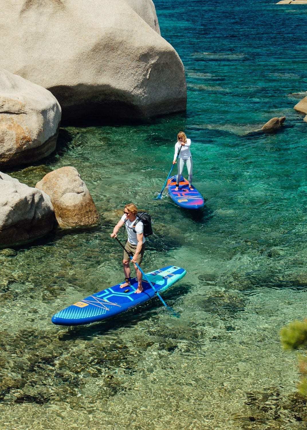 Two paddleboarders on blue boards navigate crystal-clear waters near large white rocks.