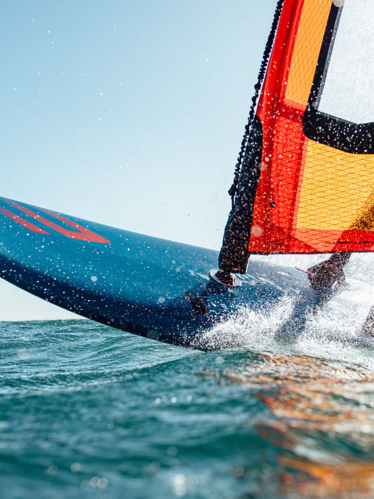 A windsurfer glides across choppy turquoise waters on a cloudy day, riding a pink and blue board with a orange and black sail 
