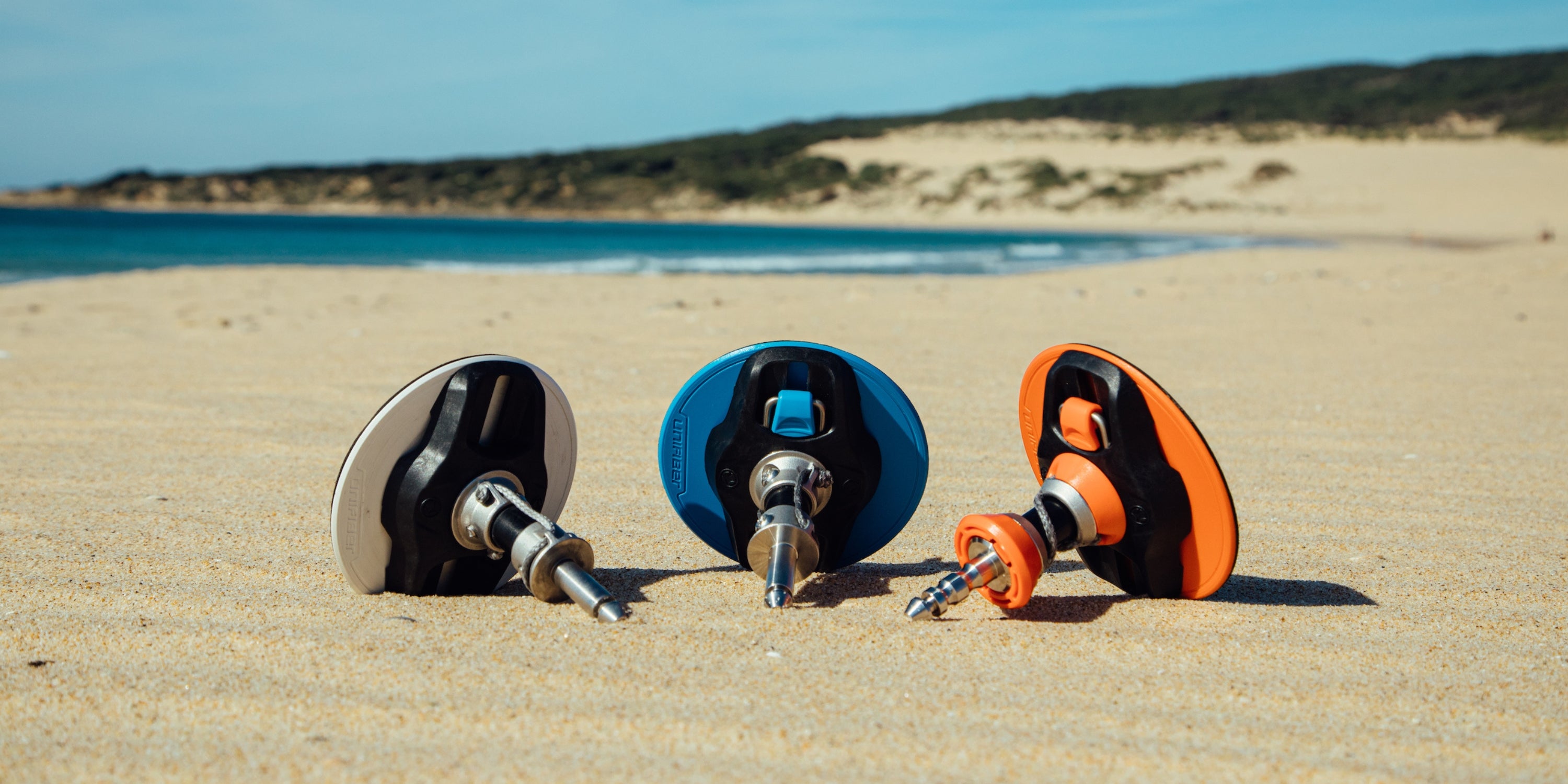 three windsurfing baseplate laid out on a sandy beach. These are the mechanisms that attach a windsurfing board to the sail 