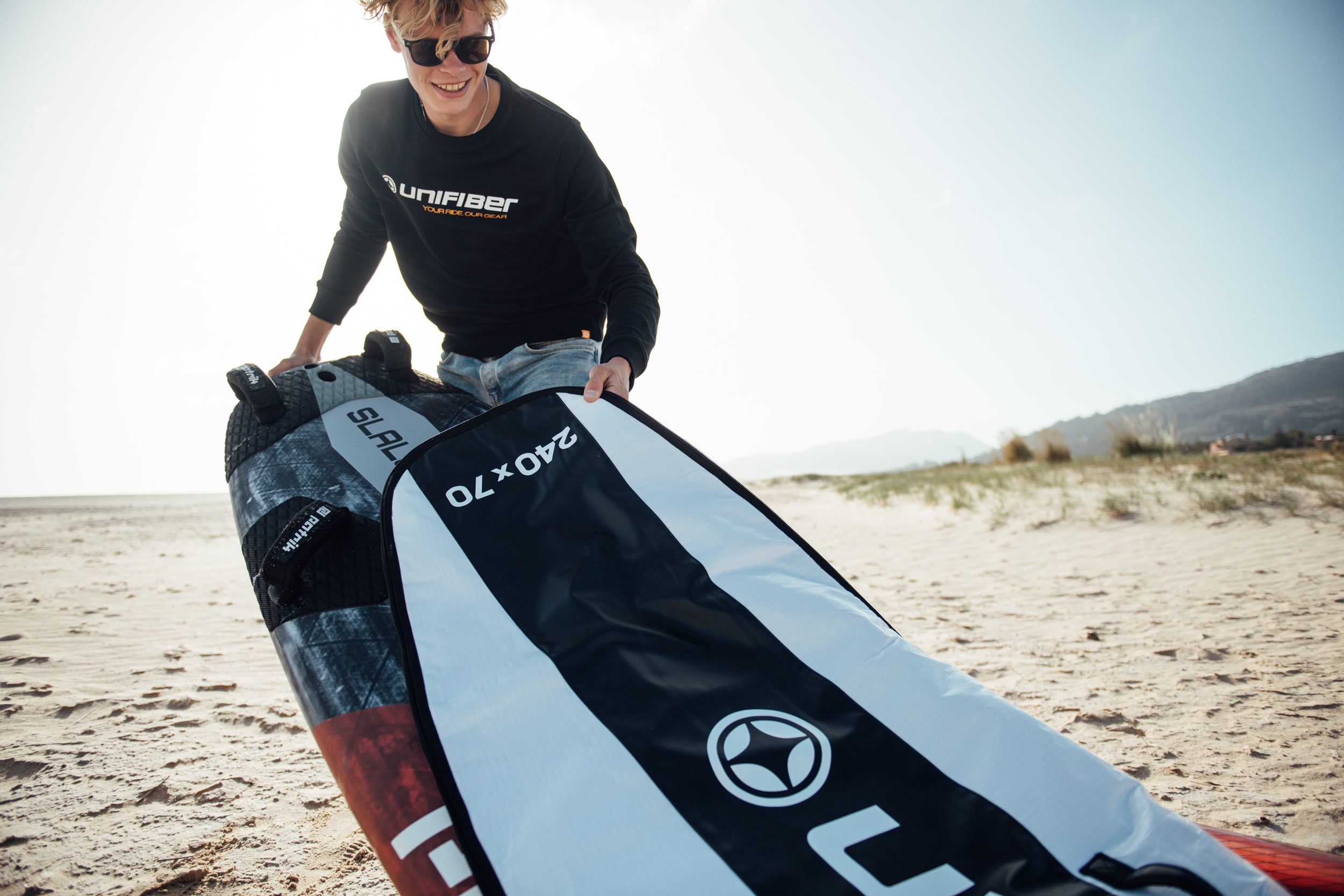 Smiling person wearing sunglasses and a black Unifiber sweatshirt demonstrating a 240x70 gear bag on a sandy beach, with dunes visible in the background