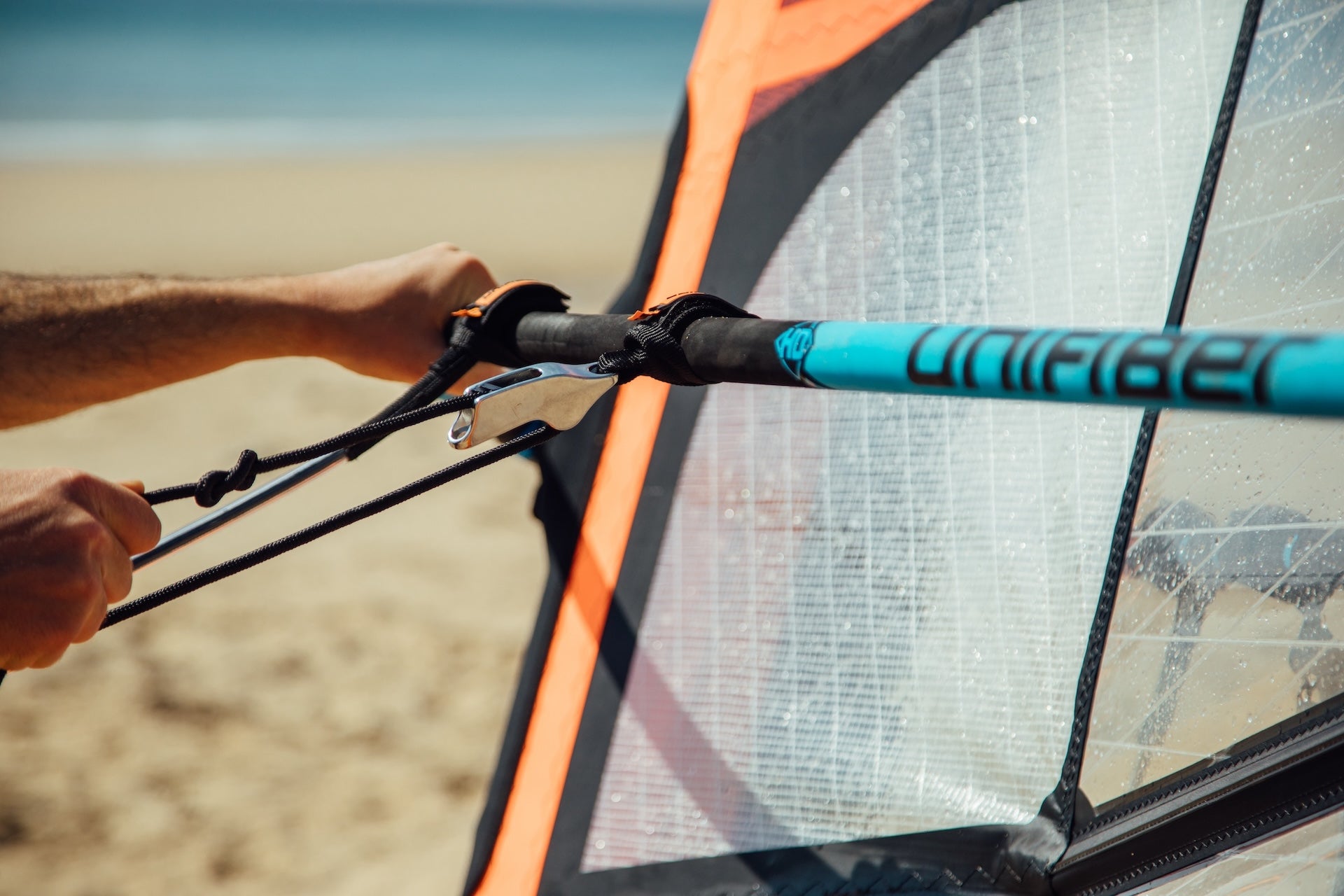 The image shows a close-up of a windsurfing rig, specifically focusing on a hand using a tool to adjust or attach rigging components. There's a boom (the black horizontal tube) with what appears to be a Unifiber mast (the light blue vertical tube with "UNIFIBER" printed on it).
