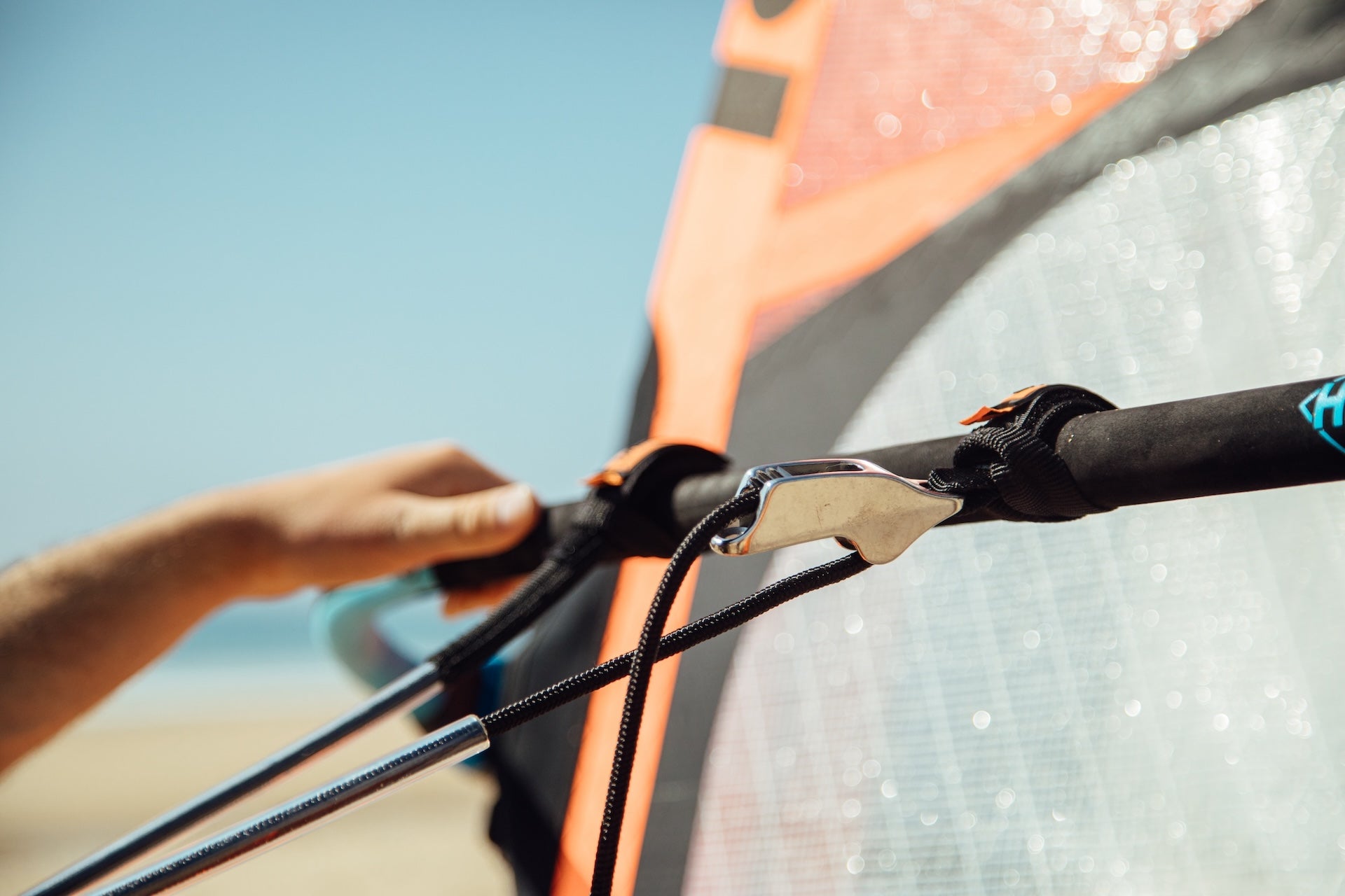 The image shows a close-up of a windsurfing rig, specifically focusing on a hand using a tool to adjust or attach rigging components. There's a boom (the black horizontal tube) with what appears to be a Unifiber mast (the light blue vertical tube with "UNIFIBER" printed on it).
