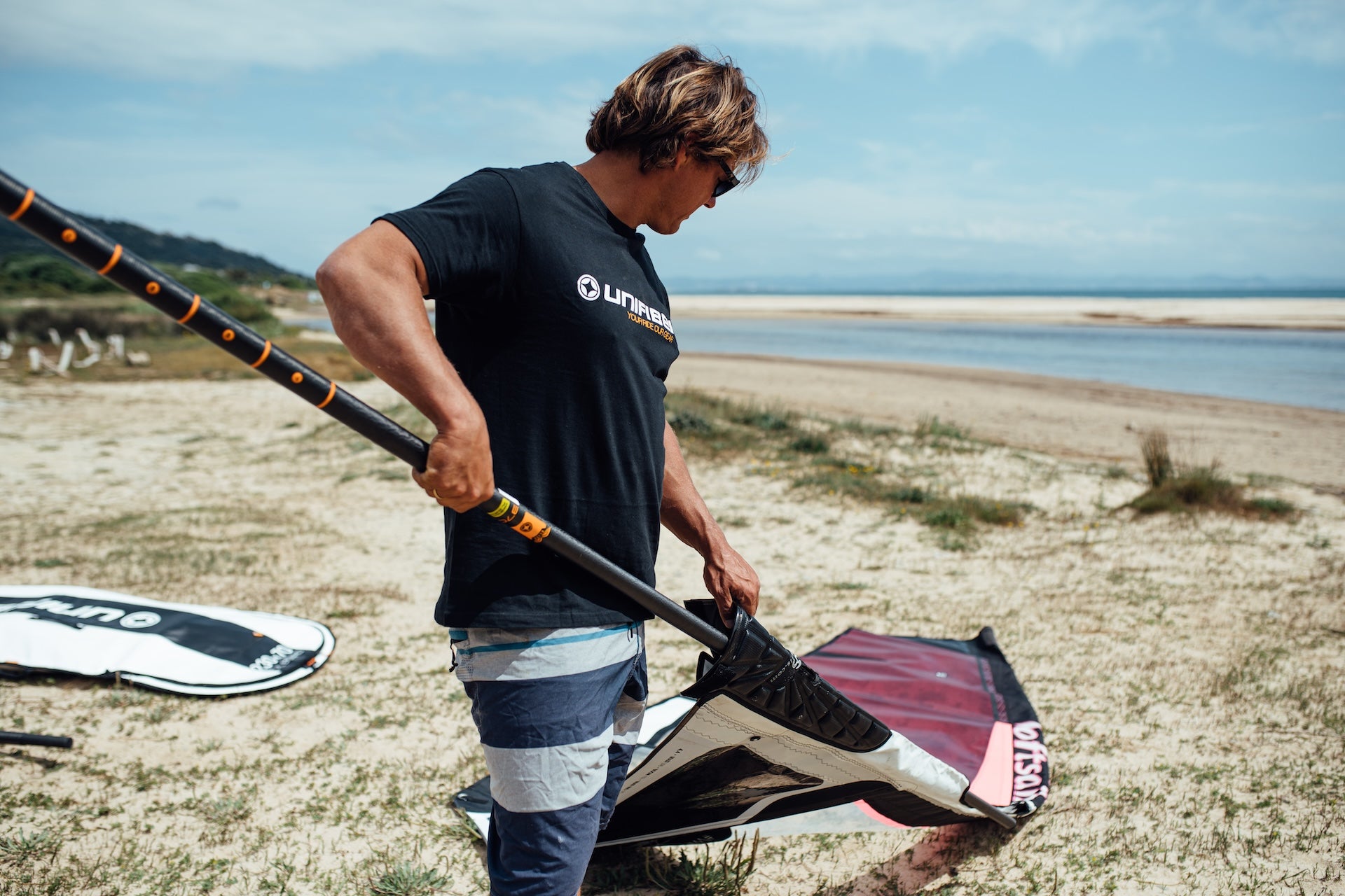 a windsurfer rigging a wave sail on the beach. He's wearing a black Unifiber t shirt and the mast have orange stripe 