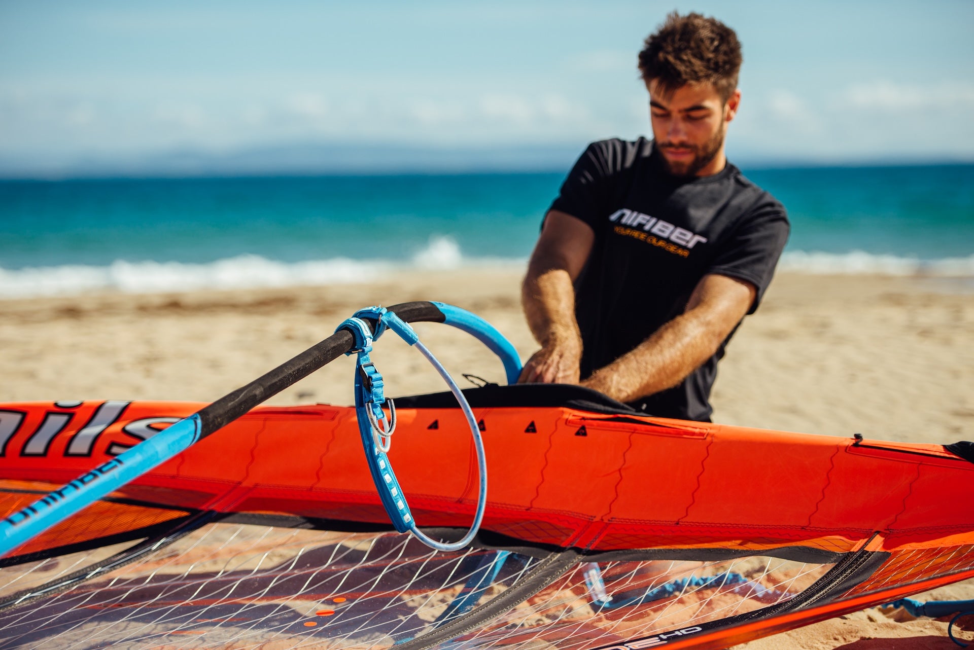 This image shows a person setting up windsurfing equipment on the beach. They're handling a orange and black sail with a Unifiber boom. The individual is wearing a black shirt, cap, and light-colored shorts against a clear blue sky backdrop with water visible in the background.