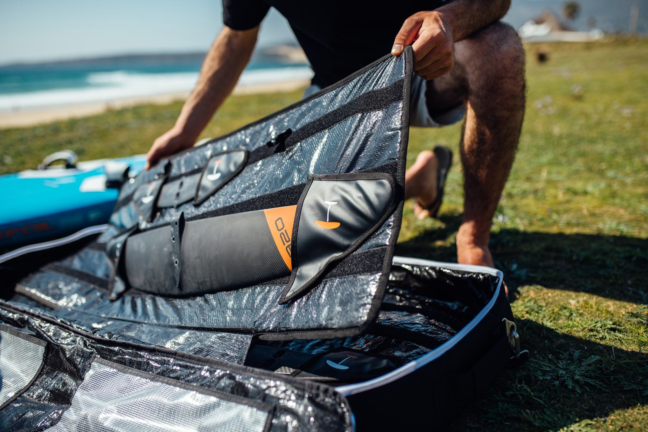 Detailed close-up of a Pro foil gear bag's reinforced padding and compartments, showing black and gray waterproof material with orange accents, being arranged on grass with ocean visible in background