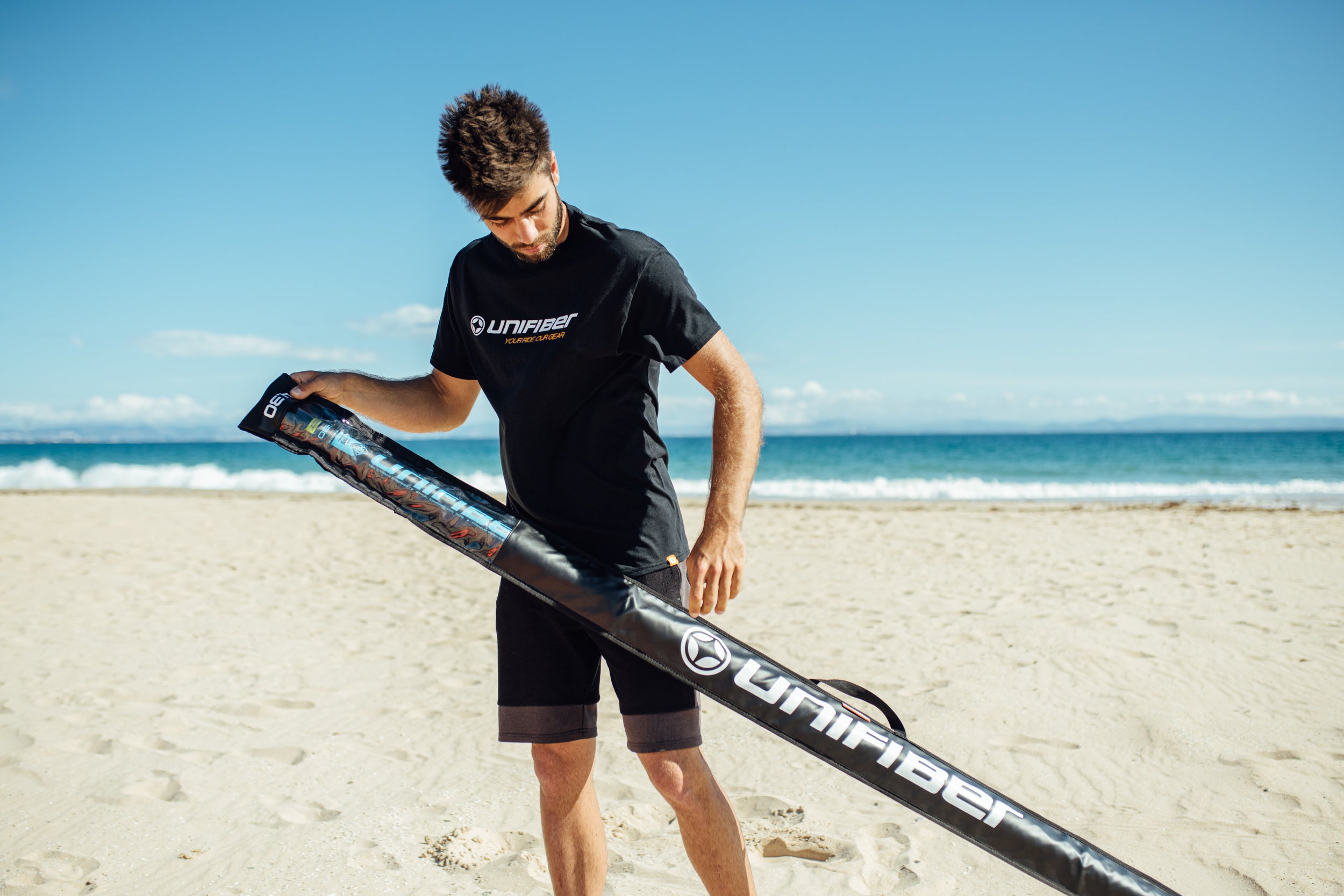 A men holding a windsurf mast on a beach with a clear Unifiber logo on hos t shirt and on the mast