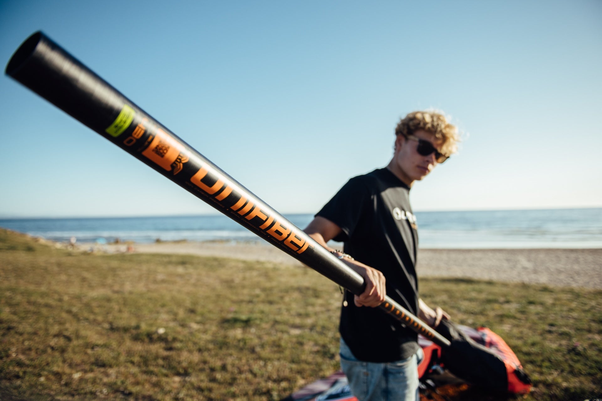 a windsurfer rigging a wave sail on the beach. He's wearing a black Unifiber t shirt and the mast have orange stripe and orange unifiber logo. 