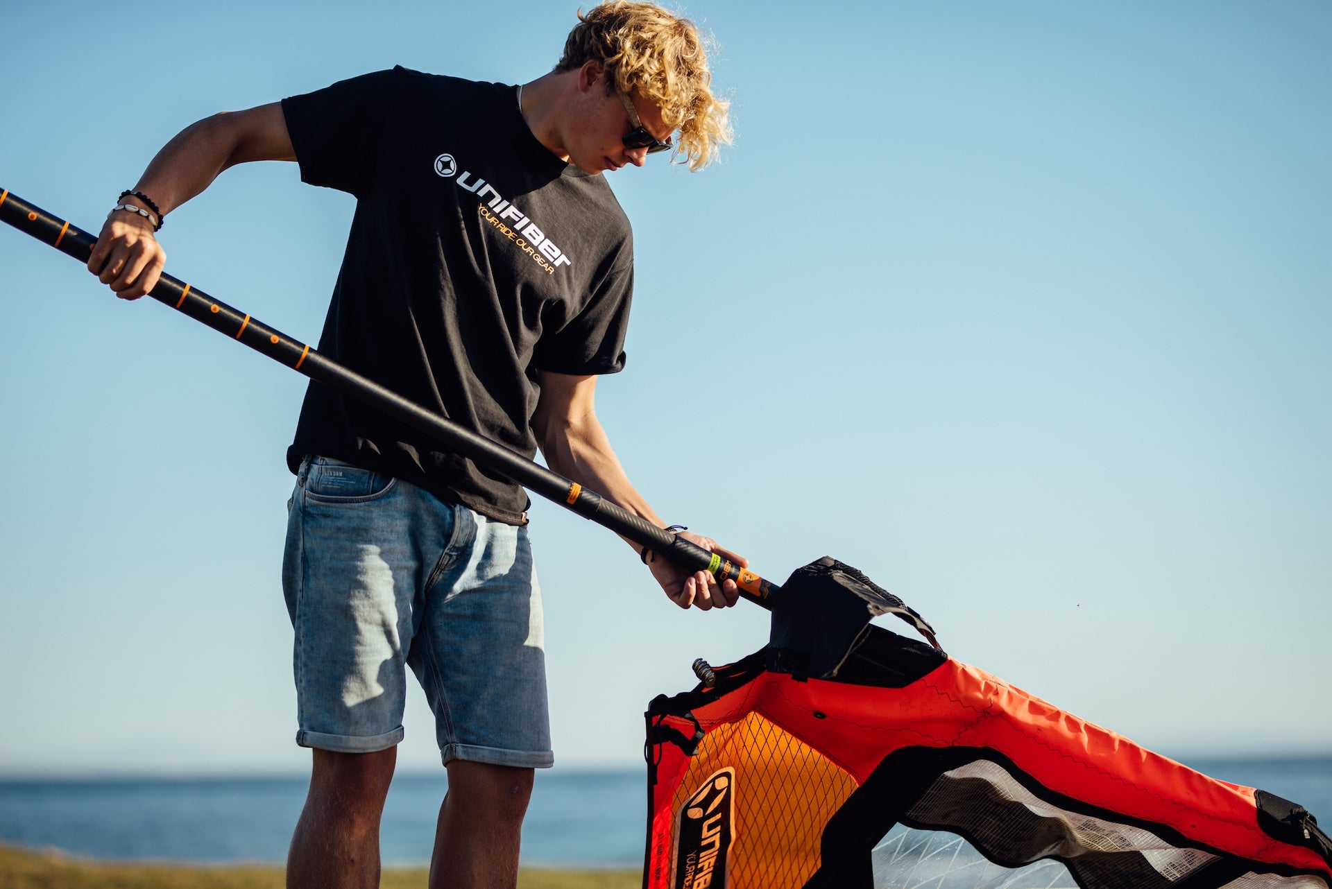 This image shows a person setting up windsurfing equipment on the beach. They're handling a orange and black sail with a Unifiber mast. The individual is wearing a black shirt, cap, and light-colored shorts against a clear blue sky backdrop with water visible in the background.