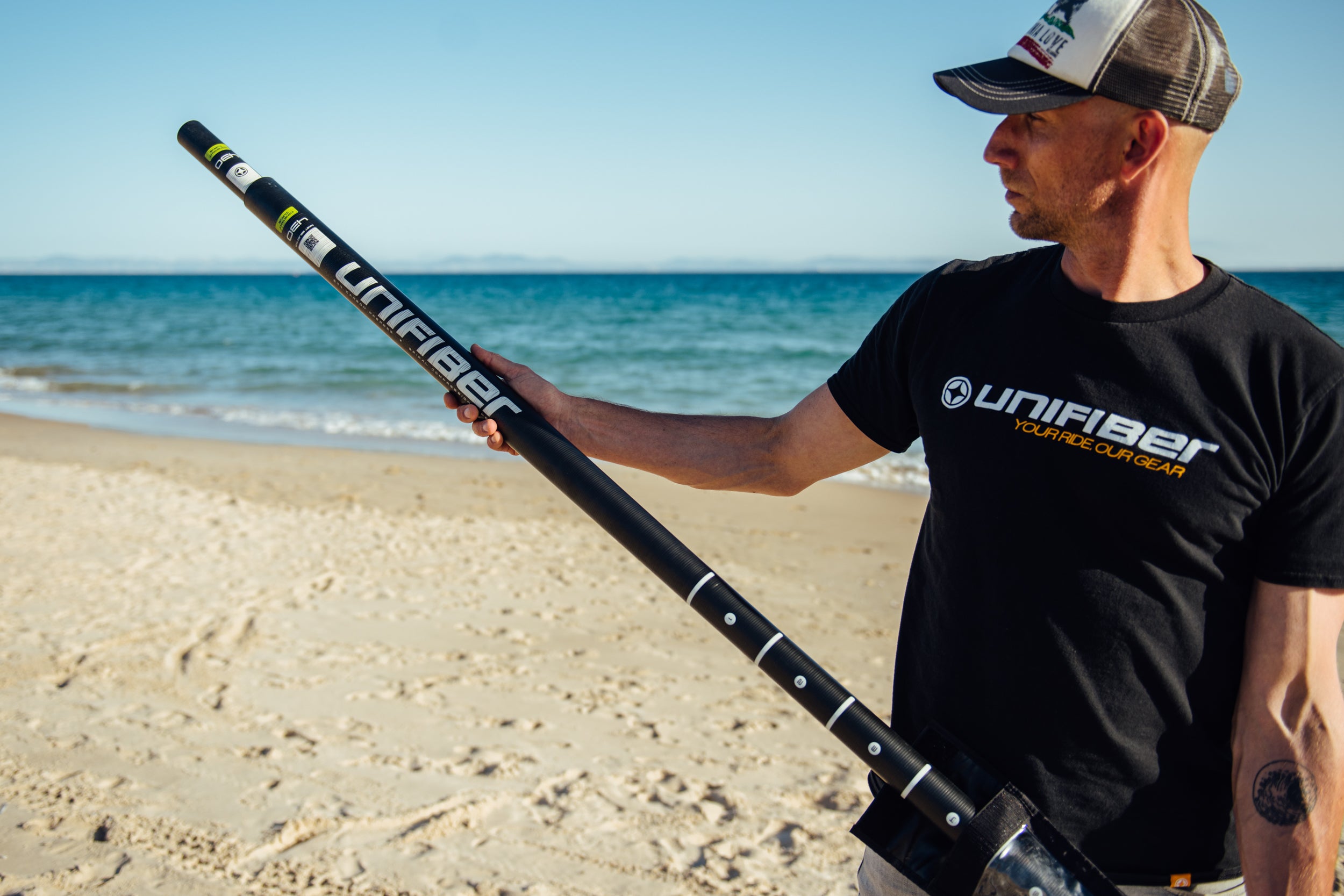 This image shows a person setting up windsurfing equipment on the beach. They're handling a mast. The individual is wearing a black shirt, cap, and light-colored shorts against a clear blue sky backdrop with water visible in the background.
