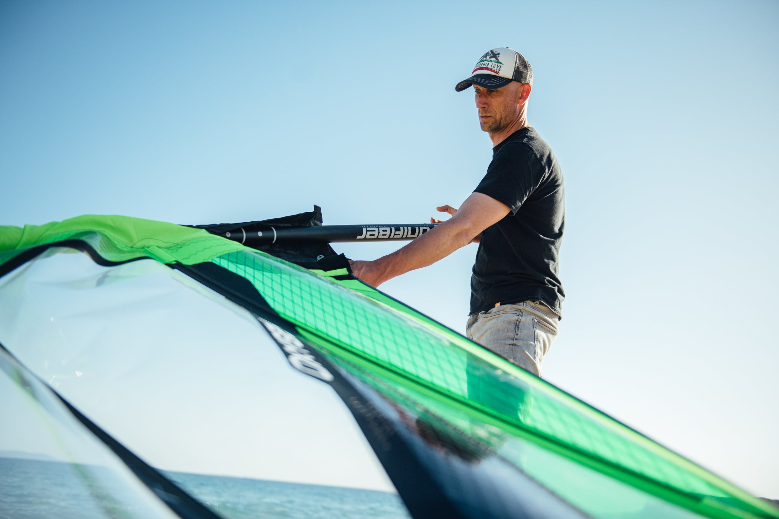 This image shows a person setting up windsurfing equipment on the beach. They're handling a green and white sail with a Unifiber boom. The individual is wearing a black shirt, cap, and light-colored shorts against a clear blue sky backdrop with water visible in the background.RéessayerClaude peut faire des erreurs. Assurez-vous de vérifier ses réponses.